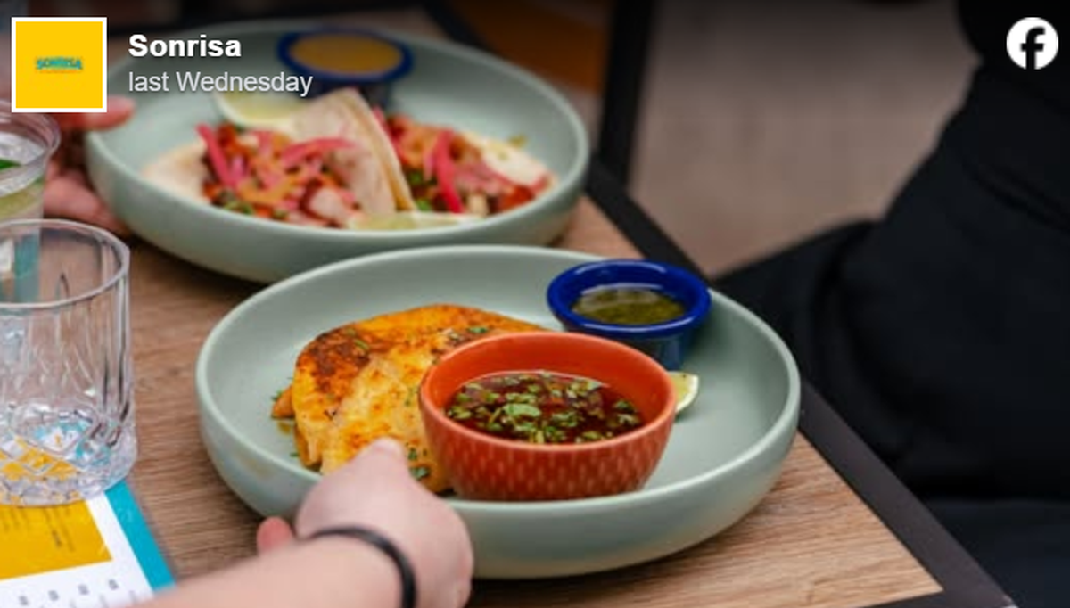 A close-up shot features two light-green plates of Mexican food served on a wooden table, with a person’s hand reaching towards the foreground dish. The front plate holds crispy, golden-brown quesadillas or birria tacos, accompanied by a small blue bowl of green salsa and a larger orange bowl filled with dipping broth topped with fresh cilantro. In the blurred background, another plate showcases tacos generously garnished with pickled red onions and fresh herbs.