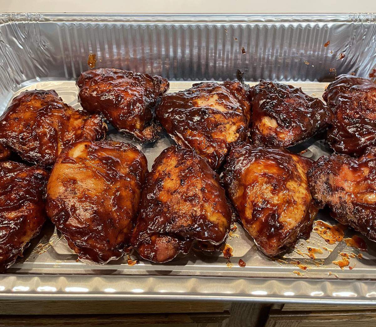 A close-up shot shows a tray of perfectly cooked barbecue chicken thighs coated in a rich, dark sauce, ready for serving. Above the tray, a wooden cutting board with the name “WOLFF” and a small wooden “BBQ” sign are visible, indicating the name of the cook or caterer.
