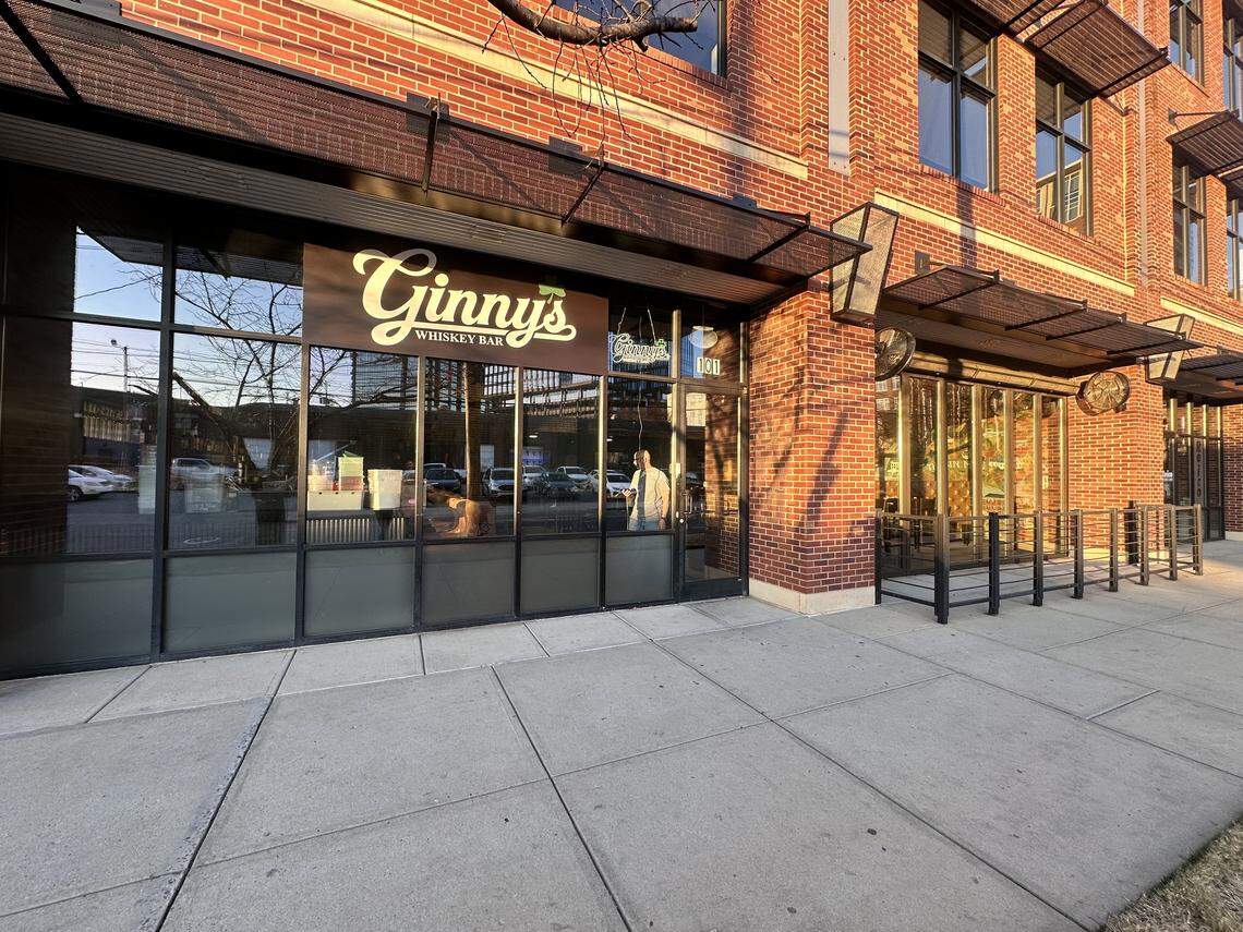 The storefront of Ginny’s Whiskey Bar features large glass windows and doors set into a red brick building with a concrete sidewalk out front. A black metal awning hangs over the entrance, and the bar’s name is displayed in a vintage-style script on a brown sign.