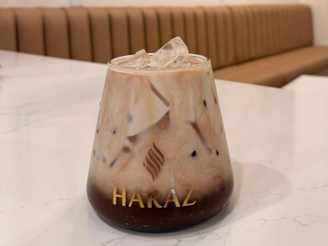 An iced coffee beverage with distinct layers of dark coffee and milk is presented in a tapered glass on a white marble table. The background is a coffee shop interior featuring a long, tan upholstered bench and a large wooden world map with the word “YEMEN” prominent at the top.