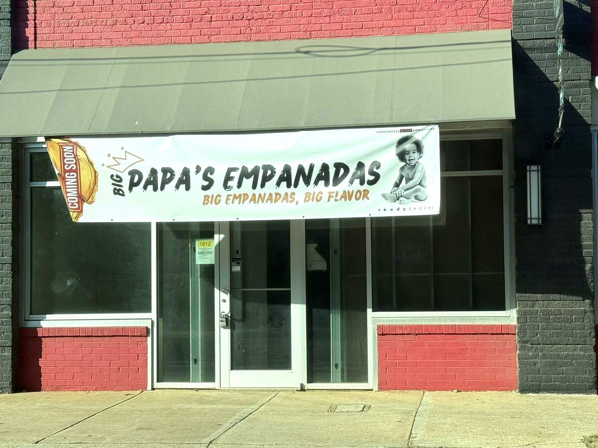 A storefront features a large white banner announcing “Big Papa’s Empanadas” with the tagline “Big Empanadas, Big Flavor.” The banner includes a “Coming Soon” graphic shaped like an empanada on the left and a black-and-white picture of a smiling baby on the right. The building itself has large glass windows and doors, set against a backdrop of red and black brick under a tan awning.