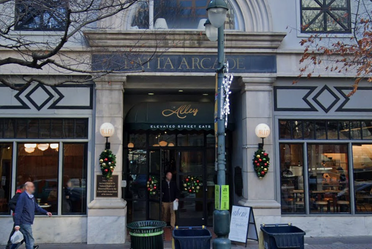The exterior entrance of the historic Latta Arcade features a grand stone facade with festive holiday wreaths and a dark green awning for “The Alley.” A few pedestrians walk along the sidewalk in front of the building’s large glass windows and classic architecture.