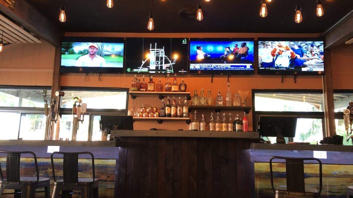 The interior view of a restaurant, showing a rustic wooden bar with black stools, several TVs playing sports, and industrial Edison bulb lighting hanging from the ceiling.