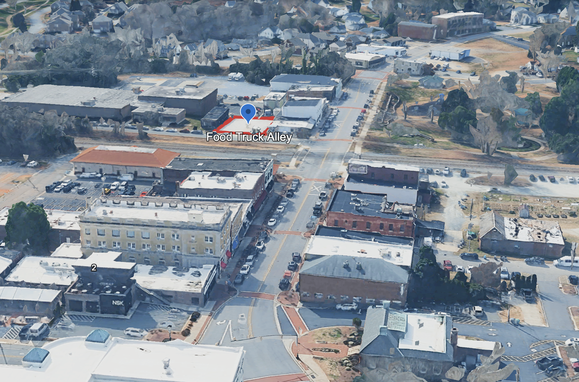 This is an angled aerial view, likely from a drone or satellite, of a small town’s downtown area. A red outline and a blue location pin mark the location of a spot labeled “Food Truck Alley” between two commercial buildings near a street intersection.