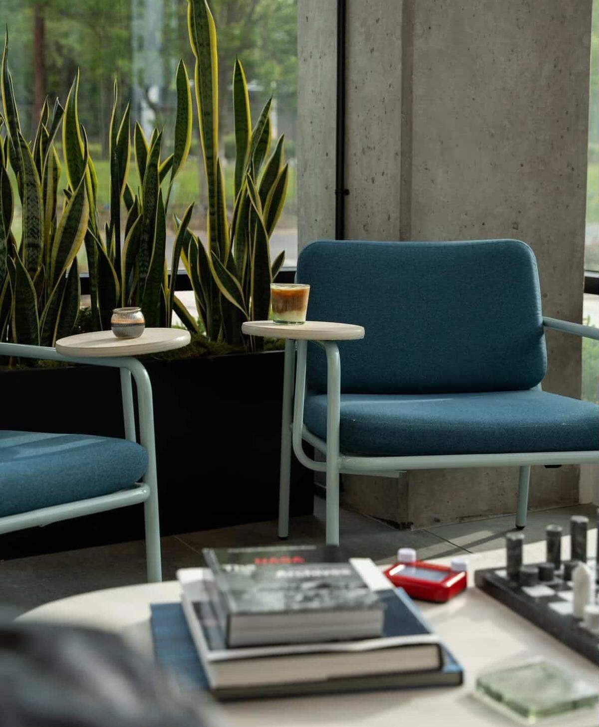 This eye-level shot features a modern teal armchair with a built-in circular side table holding a layered iced coffee. In the background, tall snake plants in a black planter sit against a concrete pillar and large windows. The foreground is slightly blurred, showing a white coffee table topped with books and a chessboard.