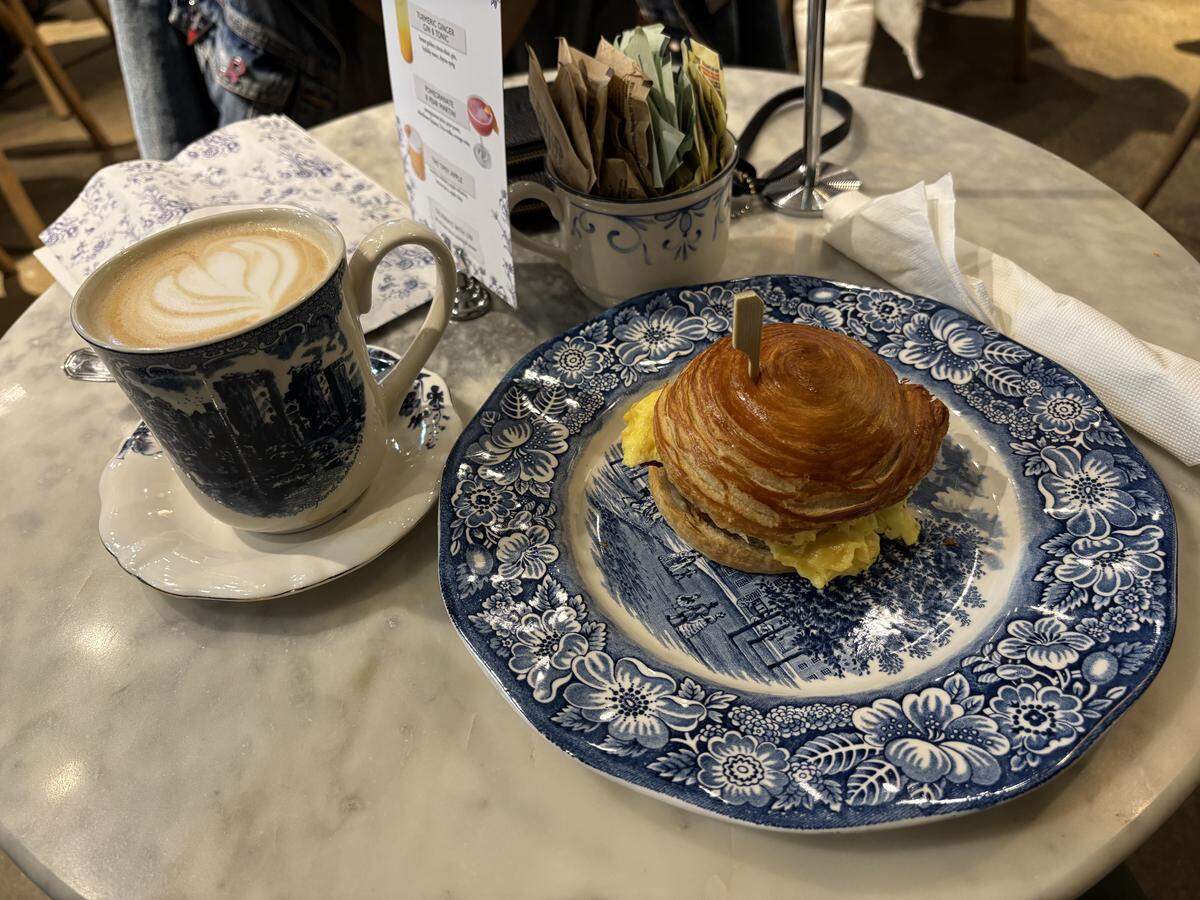 A latte with heart-shaped foam sits in a blue and white patterned mug next to a breakfast sandwich on a matching plate. The sandwich features scrambled eggs tucked inside a flaky, spiraled pastry roll on a marble tabletop.