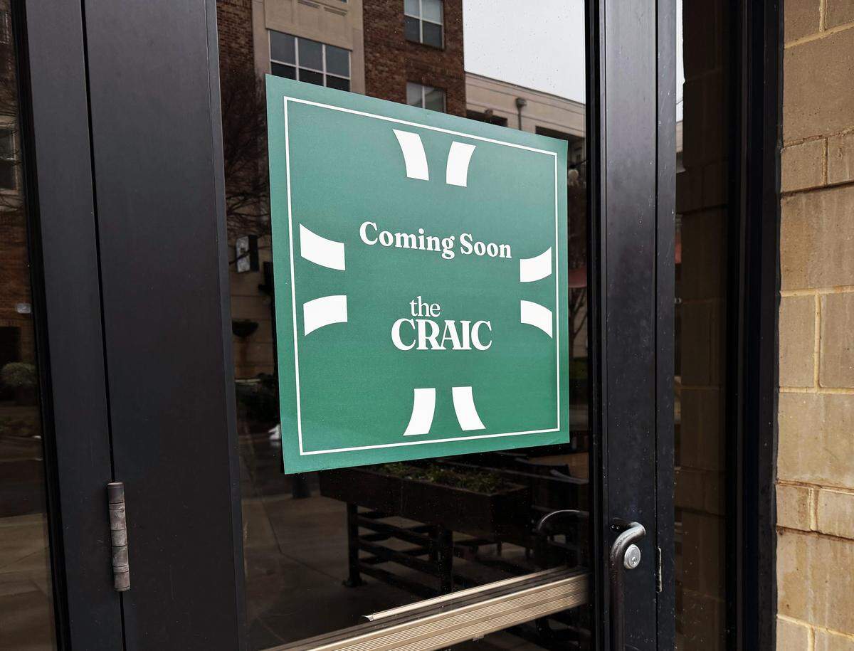 A green square sign is posted on a glass door, prominently displaying the words “Coming Soon” above the name “the CRAIC” in white text. The sign is bordered by a white frame with stylized geometric accents at the top, bottom, and sides.