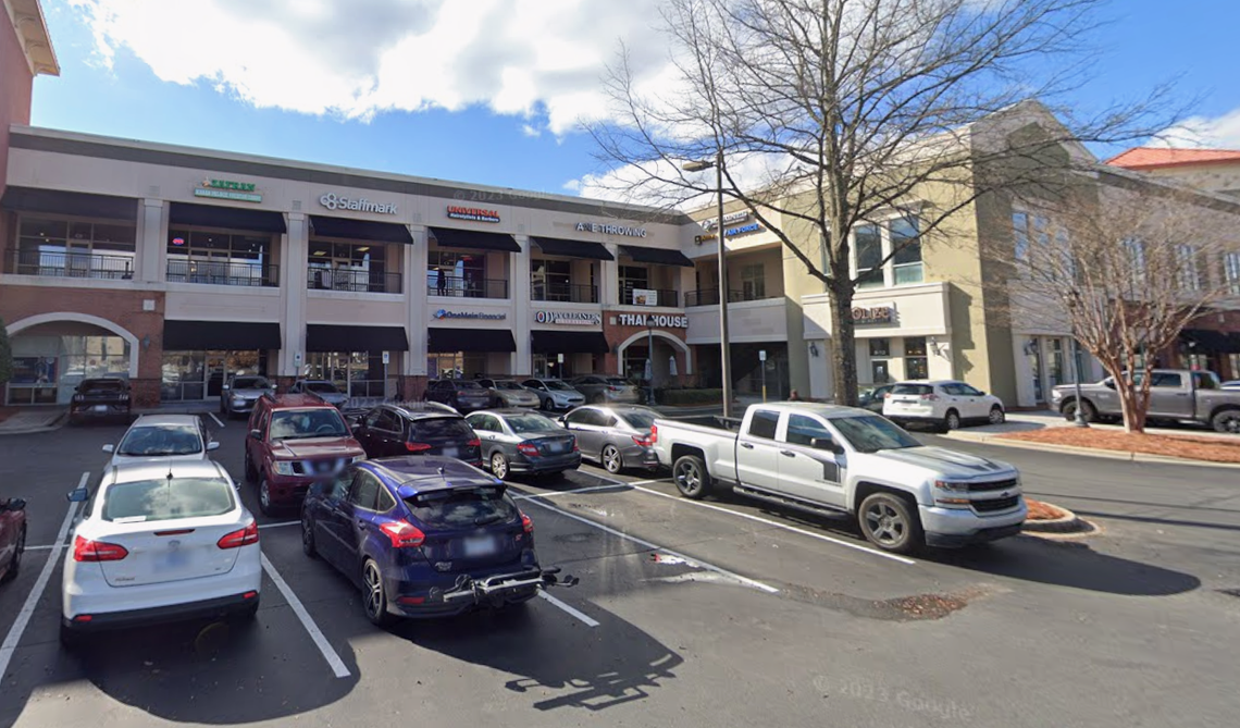A wide shot shows a two-story outdoor shopping center with several businesses, including Thai House, Staffmark, and OneMain Financial. Various cars and a silver pickup truck are parked in the asphalt lot in the foreground under a clear blue sky.