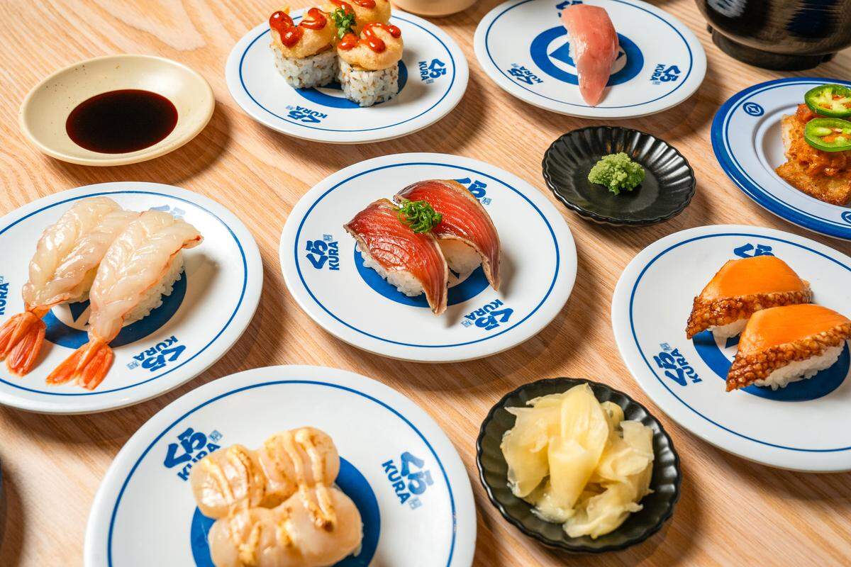 A high-angle shot displays an array of sushi plates arranged on a light-colored wooden table, featuring variety such as raw shrimp, seared scallops, and glazed eel. Small black dipping dishes containing pickled ginger, wasabi, and soy sauce are interspersed between the white and blue plates. The bright, even lighting emphasizes the vibrant colors and fresh presentation of the seafood.