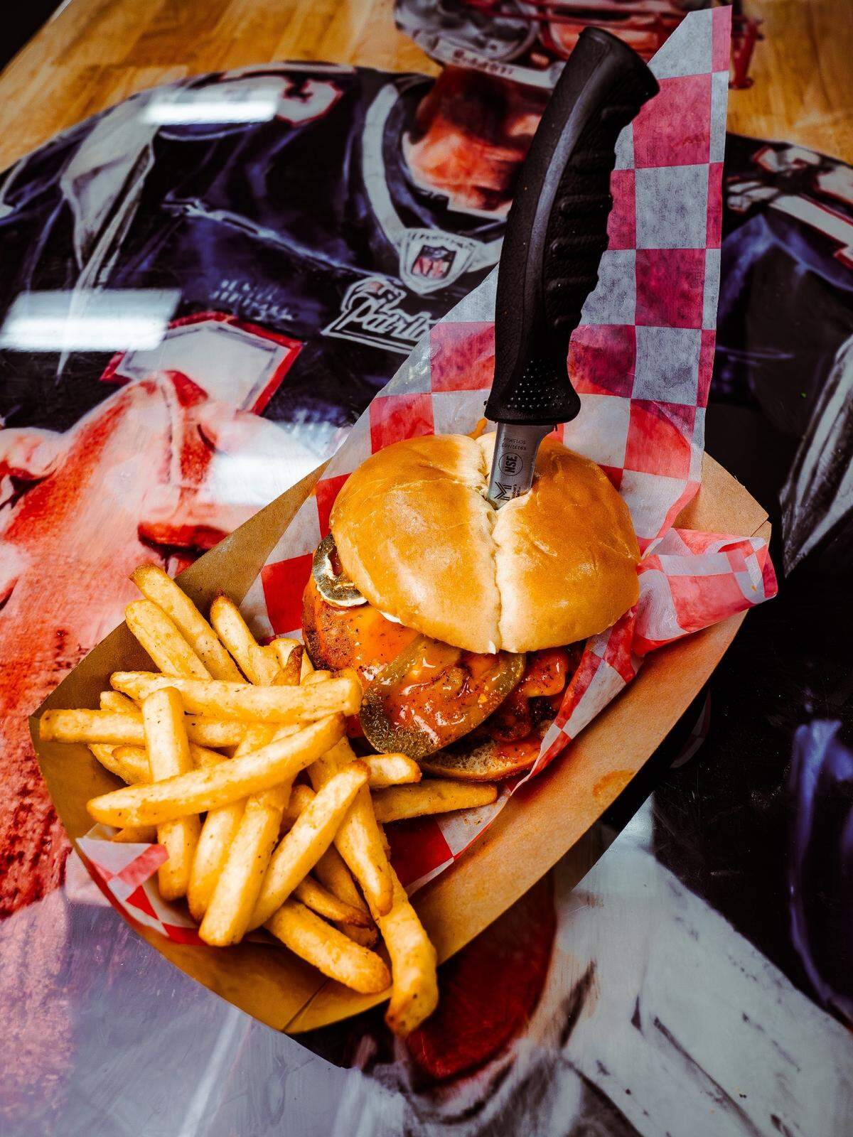 A large chicken sandwich topped with pickles and buffalo sauce is served in a paper basket alongside a generous portion of golden fries. A black-handled steak knife is pierced through the center of the bun to hold the sandwich together.