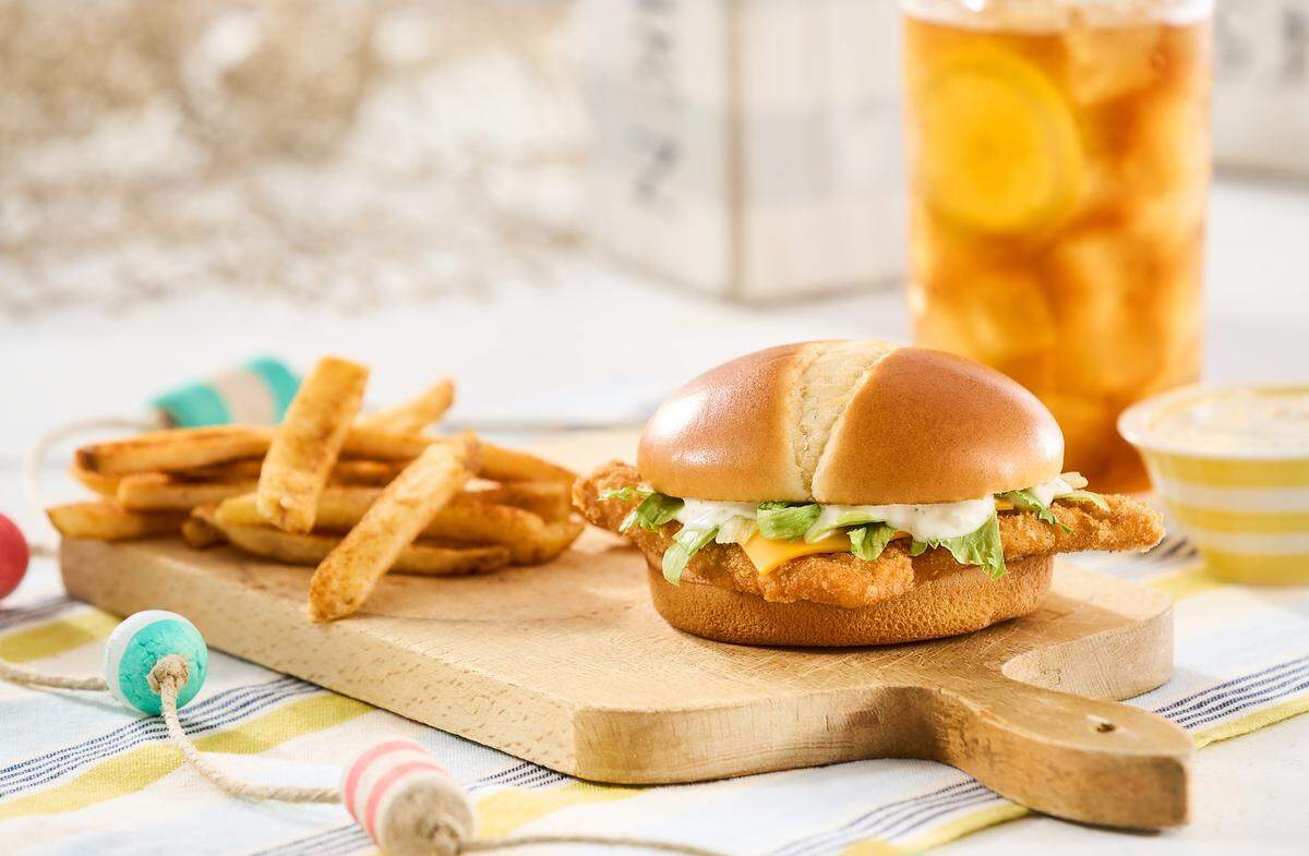 Gemini said A Bojangles Bojangler fish sandwich, featuring a breaded fillet with melted cheese, tartar sauce, and shredded lettuce on a toasted bun, is served on a wooden board alongside seasoned fries. The meal is accompanied by a large glass of iced tea with lemon in a bright, coastal-themed setting.