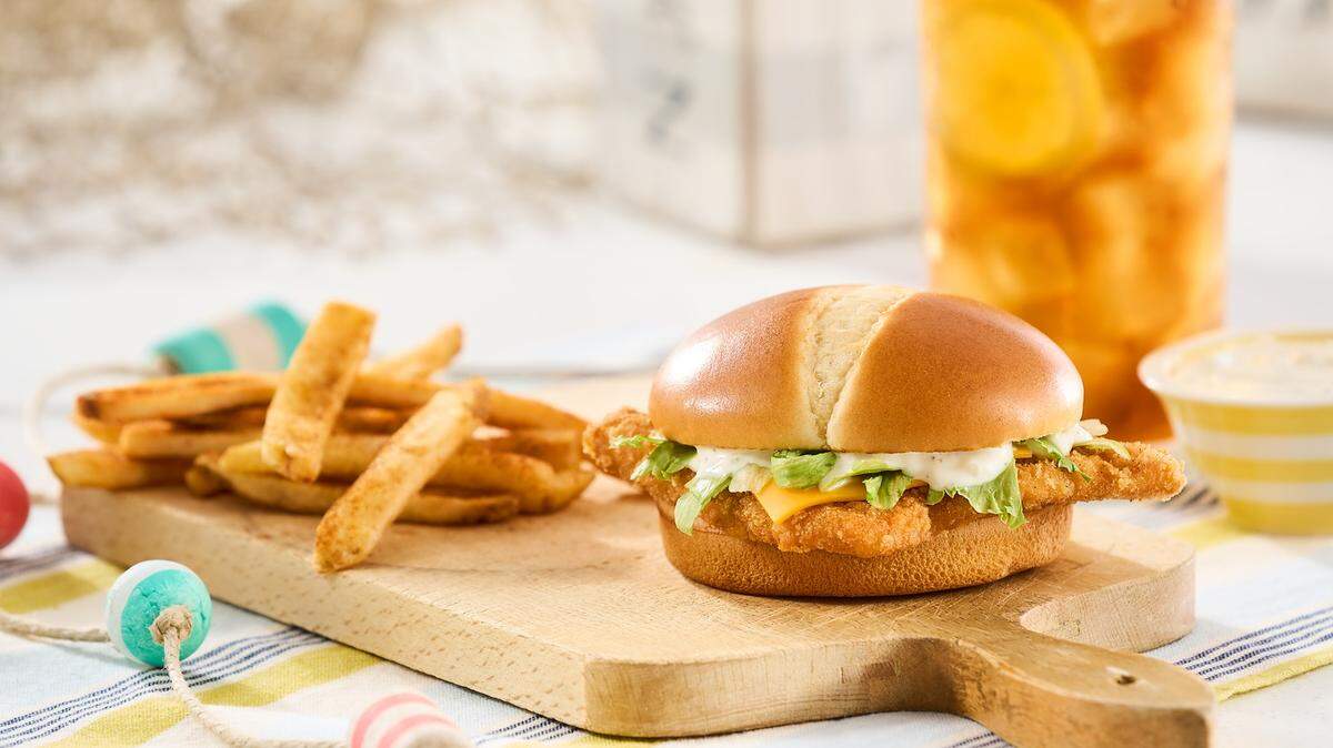 Gemini said A Bojangles Bojangler fish sandwich, featuring a breaded fillet with melted cheese, tartar sauce, and shredded lettuce on a toasted bun, is served on a wooden board alongside seasoned fries. The meal is accompanied by a large glass of iced tea with lemon in a bright, coastal-themed setting.