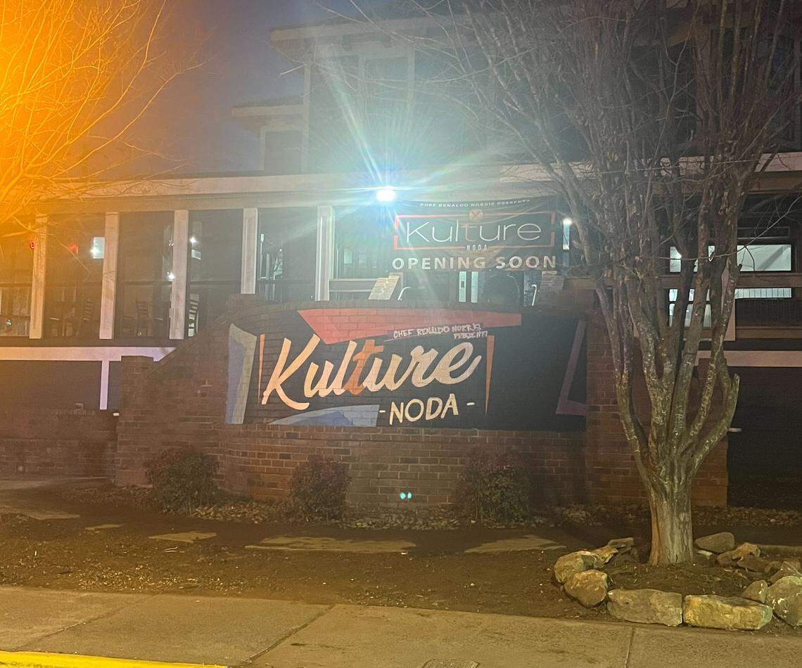 A storefront for a restaurant called “Kulture - NoDa” is shown at night with a large mural and an “Opening Soon” banner. The brick exterior features a stylized logo and mentions Chef Renaldo Norris, while a bright street lamp casts a glare over the scene.