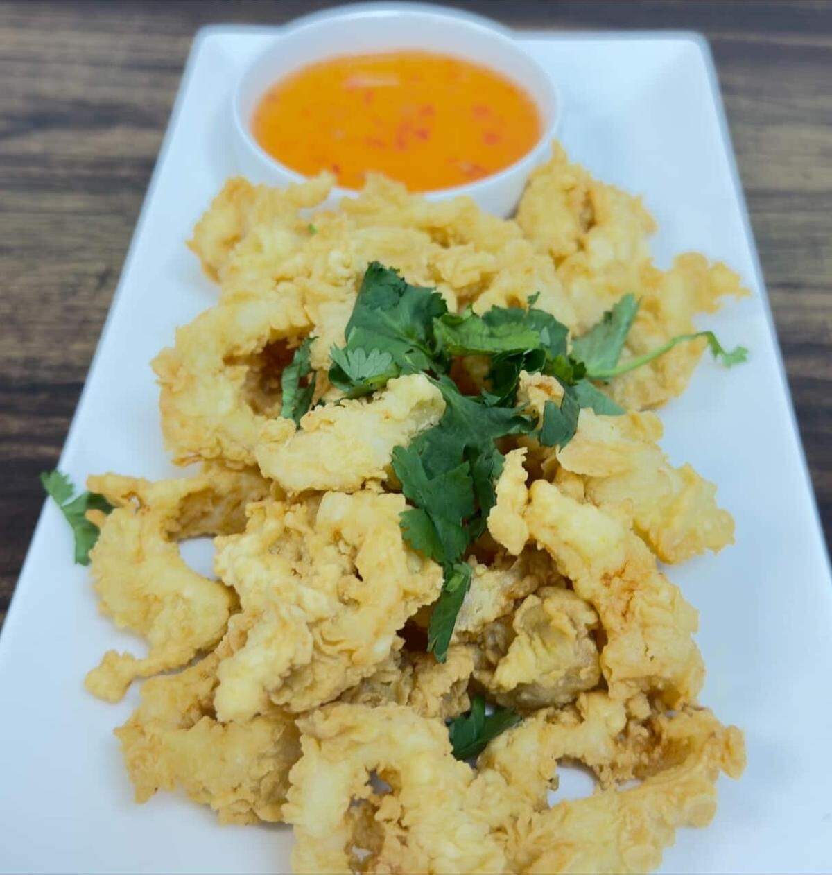A generous pile of golden-brown, battered calamari rings is garnished with fresh cilantro and served on a white rectangular plate. A small side of bright orange sweet chili dipping sauce completes the presentation.