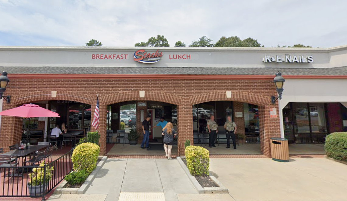 A storefront view of a restaurant called Stacks Kitchen, which serves breakfast and lunch, located in a brick strip mall next to K & E Nails. The restaurant entrance is framed by three brick arches, with several people standing near the doorway and outdoor patio seating visible to the left.