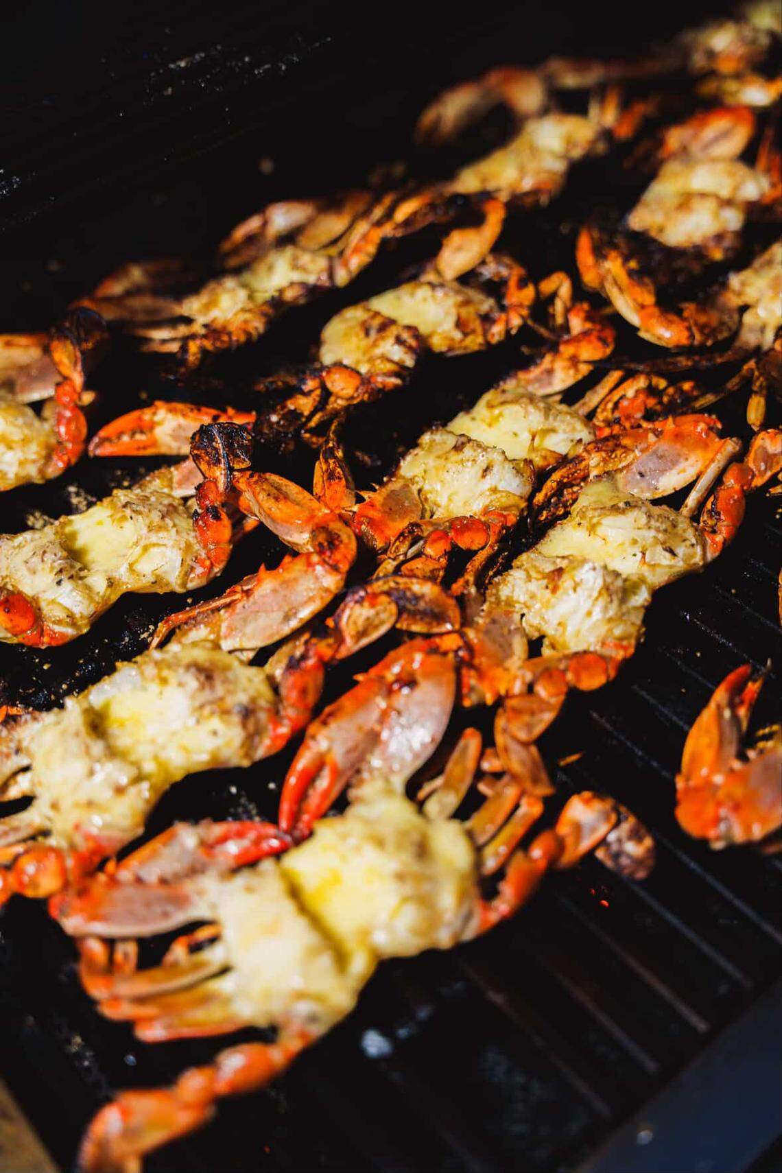 Dozens of blue crabs are lined up across a black grill, their shells turning a vibrant orange from the heat. Each crab is split open and topped with a savory, melted garlic butter seasoning. The close-up shot captures the steam and sizzle of the seafood as it cooks over the open flame.