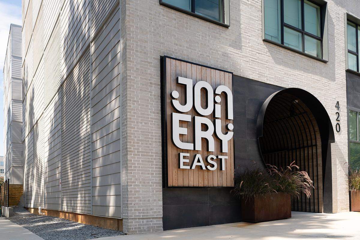 A large, modern building sign for “Joinery East” features bold white 3D lettering mounted on a vertical wood-paneled backing against a light brick wall. To the right of the sign, a striking circular arched entryway leads into the building, flanked by large rusted metal planters with tall ornamental grasses.