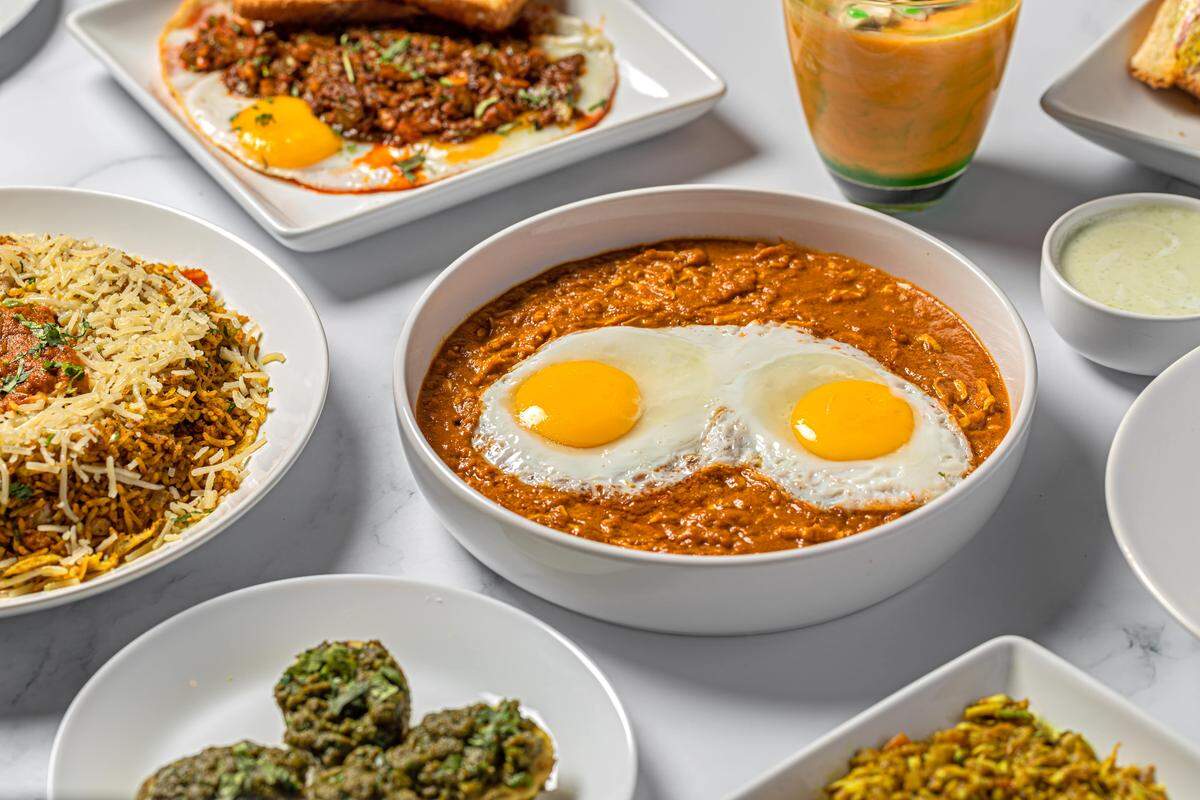A high-angle view of several Indian breakfast and lunch items, with a bowl of curry topped with two fried eggs as the central focus. Other plates include biryani, spiced ground meat with eggs and toast, and green, savory fried patties.