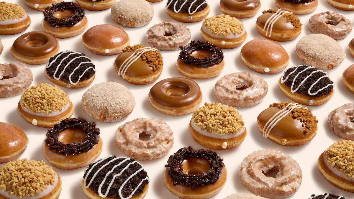 A close-up, overhead view displays a large randomized grid of various doughnuts, filling the entire frame. The assortment includes ring doughnuts, shell doughnuts, and several with decorative toppings like chocolate crumbs, cream glaze, and white stripes on a caramel base.