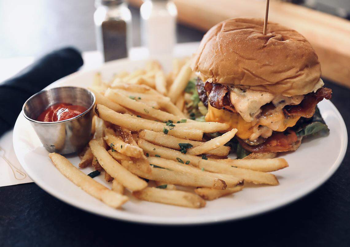A generous portion of seasoned french fries and a large bacon cheeseburger are presented on a white plate. The burger, held together by a toothpick, is topped with cheese, bacon, and sauce, and the fries are served with a small metal cup of ketchup.