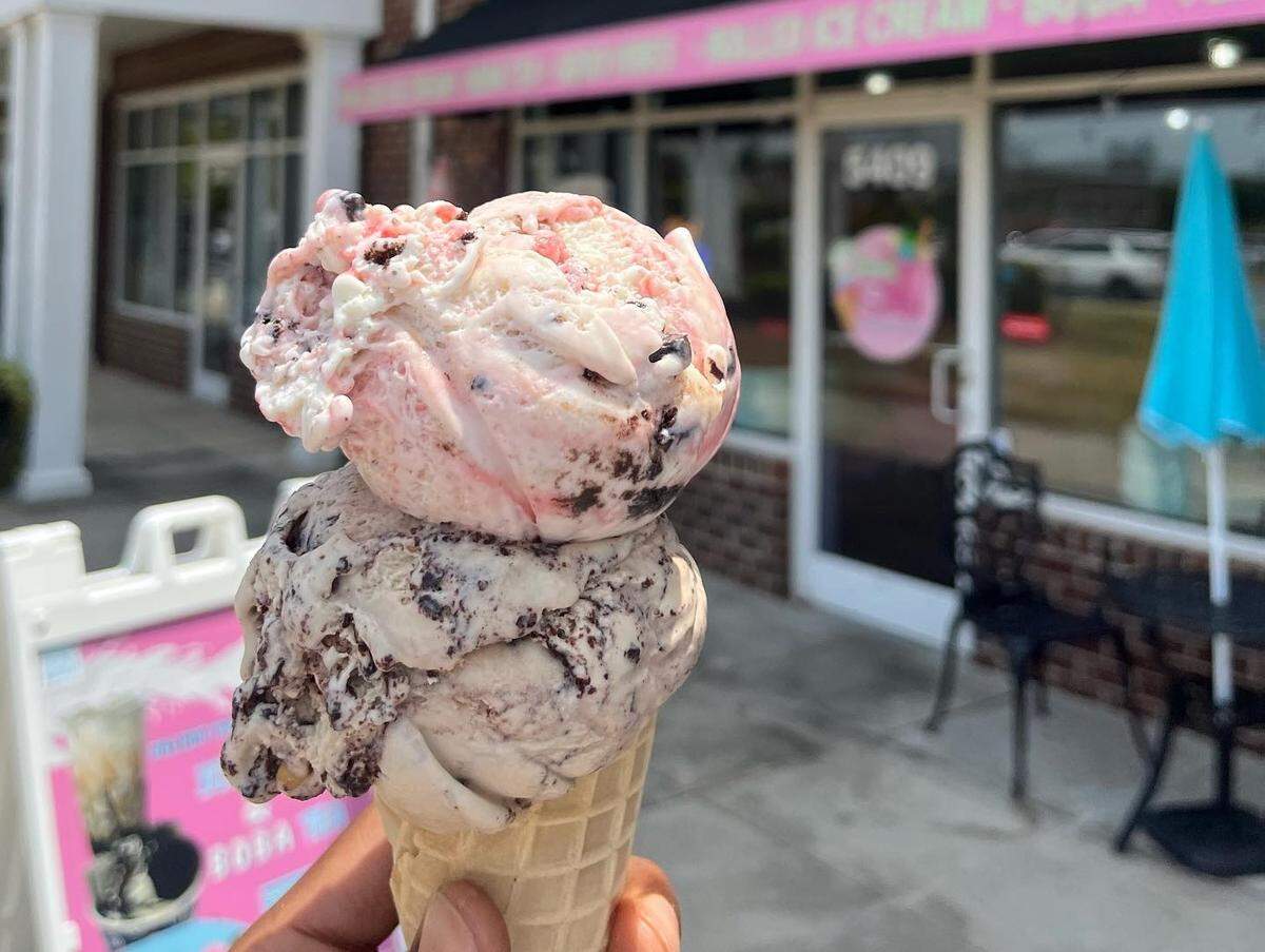 A person holds an ice cream cone with two large scoops: one is a dark chocolate chip flavor and the top scoop is a pink and white cherry flavor. The ice cream is held in front of a brick building with a pink awning that reads “ICE CREAM - BOBA.”