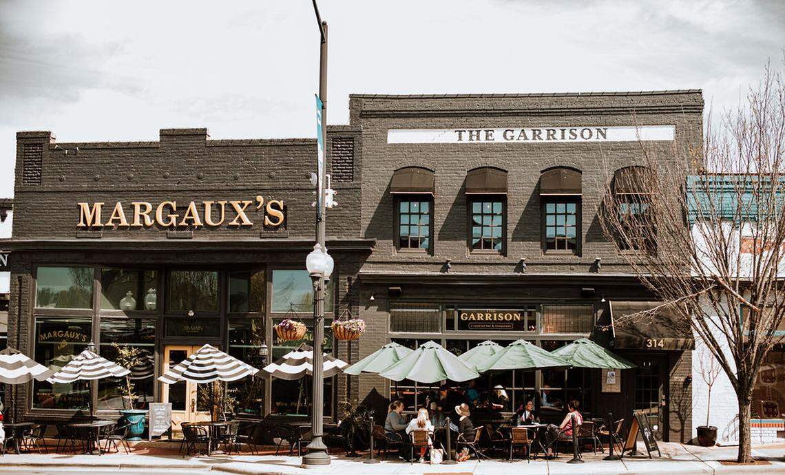 The side-by-side exteriors of Margaux’s and The Garrison restaurants are shown, featuring dark brick facades and outdoor patio seating. Patrons are visible sitting under green and striped umbrellas on the sidewalk on an overcast day.