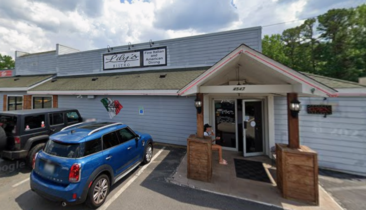 A wide shot of the exterior of Lily’s Bistro, a light gray building featuring a sign that reads “Fine Italian and American Dining.” A bright blue car is parked in the foreground near the entrance, where a person is sitting on a small porch area under a gabled roof.