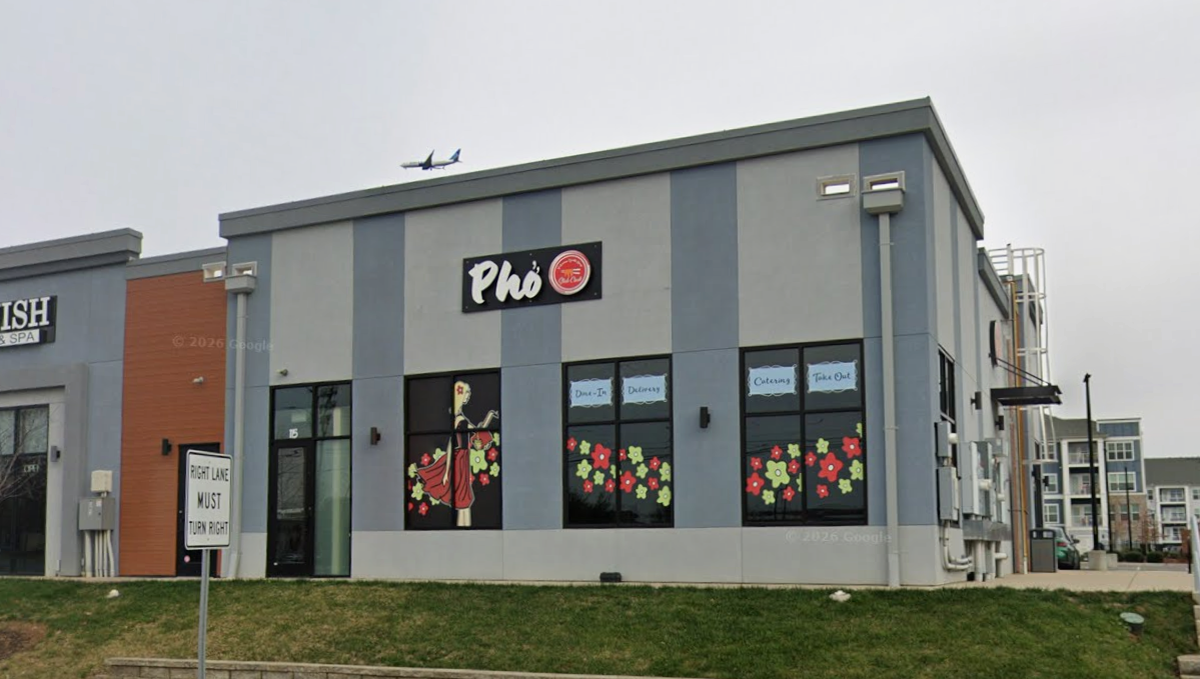 An exterior view shows a modern, grey and blue striped commercial building featuring a Vietnamese restaurant called “Phở”. The windows are decorated with colorful flower decals and a graphic of a woman in a red dress, while a “Right Lane Must Turn Right” sign stands on the grassy slope in front. In the overcast sky above the rooftop, a commercial airplane is visible in mid-flight.