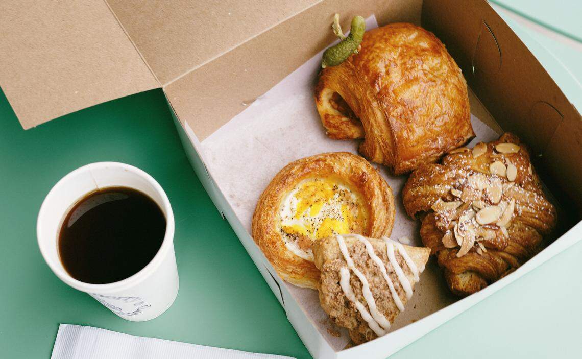 An overhead shot shows a white paper coffee cup next to a cardboard box filled with four assorted pastries on a mint green table. Inside the box, there is a savory egg-topped danish, a flaky almond croissant, a glazed scone, and a golden-brown turnover. A single triangular slice of cake topped with white frosting and edible flower petals sits on a white napkin in the foreground.