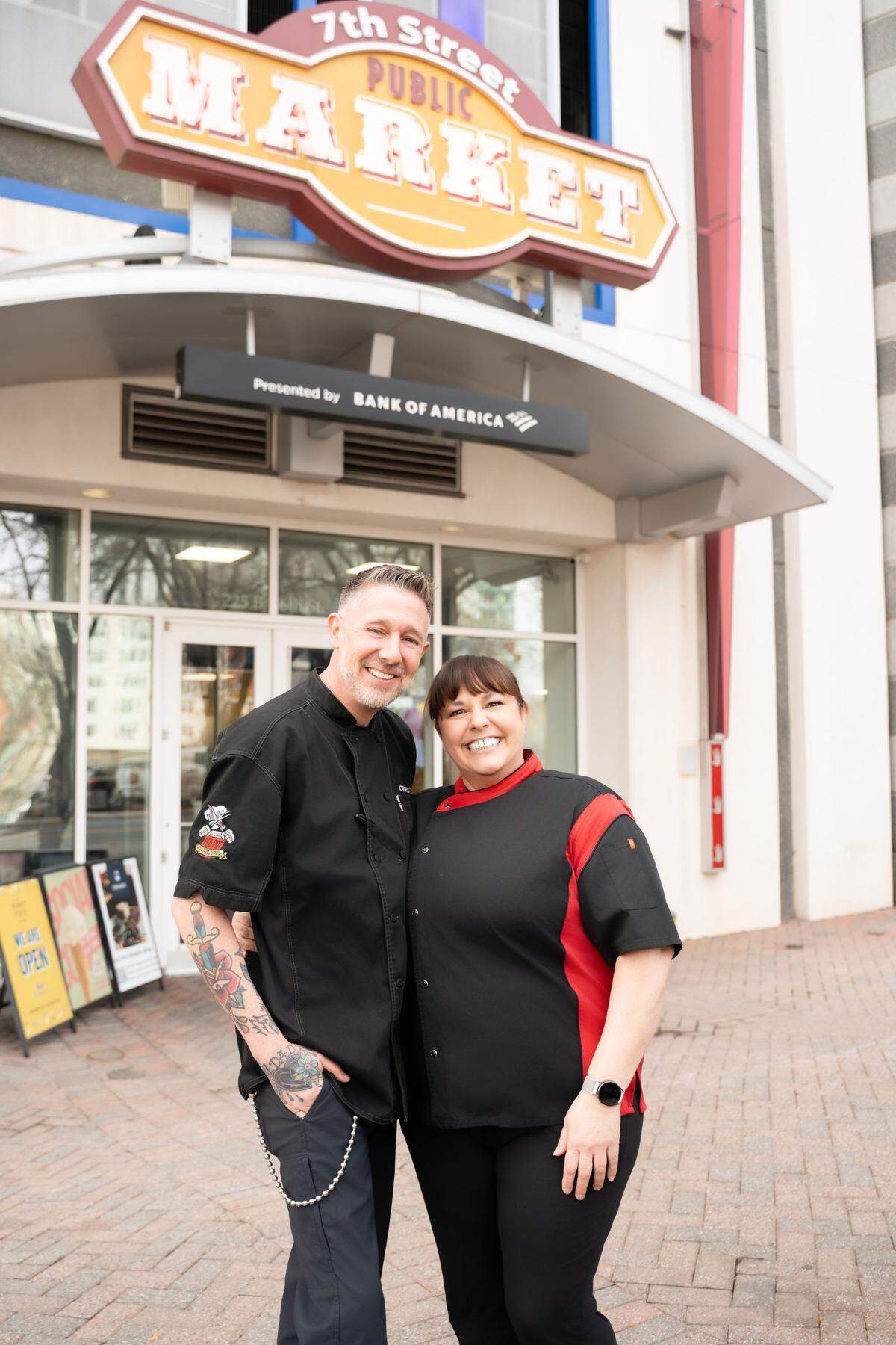 A medium shot of two cheerful chefs posing outside the 7th Street Public Market. The man on the left wears a black chef’s tunic and has visible arm tattoos; the woman on the right wears a black chef’s coat with red accents. The market’s vintage-style wooden sign hangs above them against a modern building backdrop.