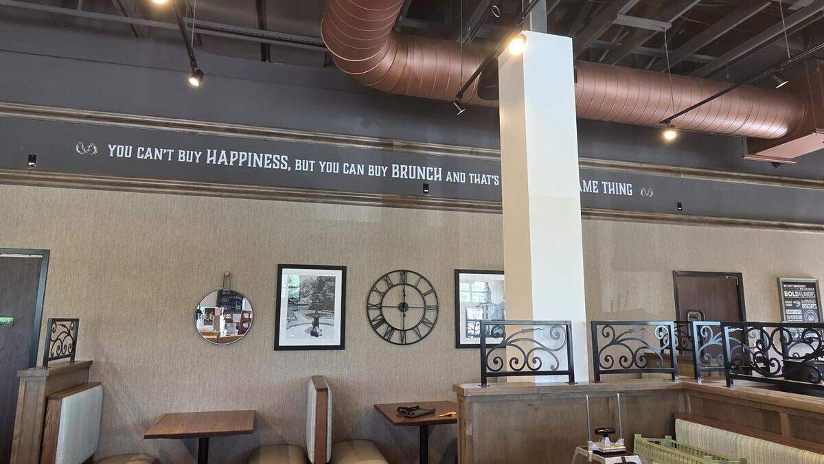 The interior of a restaurant features a wall quote that reads, “YOU CAN’T BUY HAPPINESS, BUT YOU CAN BUY BRUNCH AND THAT’S PRETTY MUCH THE SAME THING.” Below the text, the textured wall is decorated with a small round mirror, two framed black-and-white photos, and a large decorative wall clock. In the foreground, wooden booths with decorative metal railings are visible under an open ceiling with exposed copper-colored ductwork.