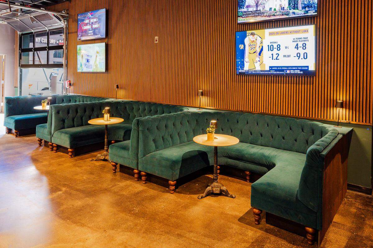 A row of curved, tufted teal velvet booths lines a wall accented with vertical wooden slats. Small, round wooden tables with individual black lamps sit in front of each booth section on a polished concrete floor. Multiple flat-screen televisions displaying sports broadcasts are mounted on the wall above the seating, and a glass garage door is visible in the background.