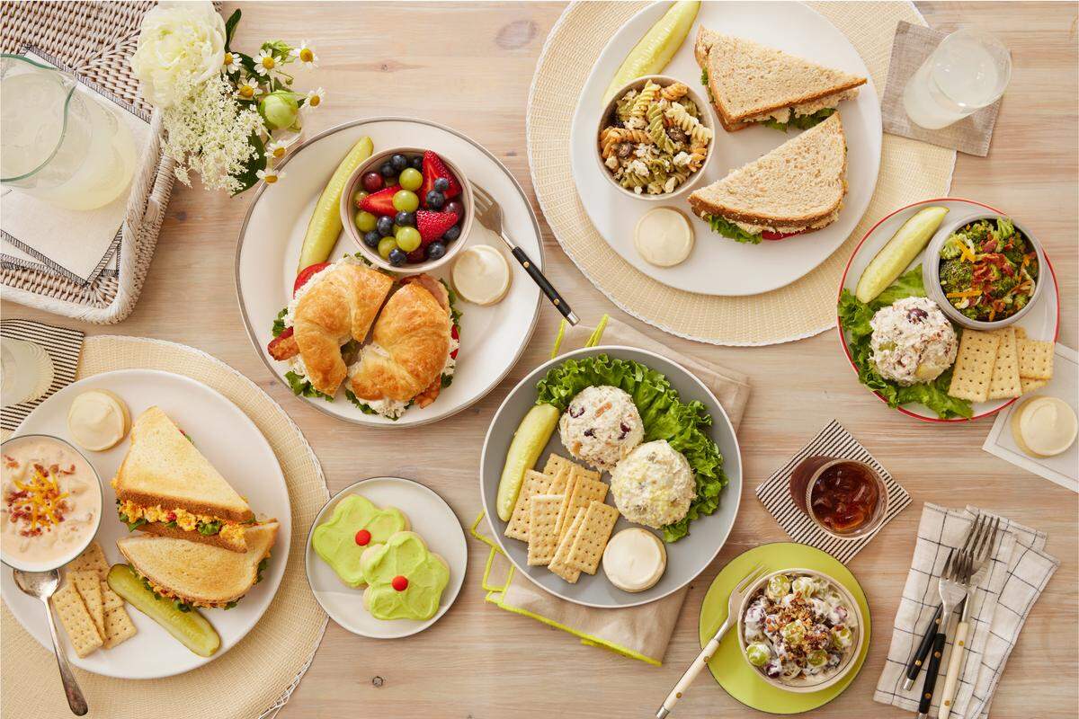 An overhead view displays a wooden table covered in various Chicken Salad Chick meals, including croissants, sandwiches, and scoops of chicken salad. The spread is accompanied by colorful sides like fruit salad, broccoli salad, and several frosted flower-shaped cookies.