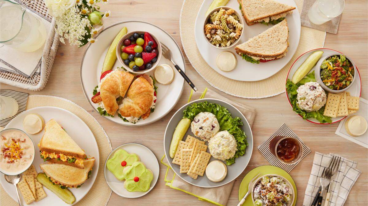 An overhead view displays a wooden table covered in various Chicken Salad Chick meals, including croissants, sandwiches, and scoops of chicken salad. The spread is accompanied by colorful sides like fruit salad, broccoli salad, and several frosted flower-shaped cookies.