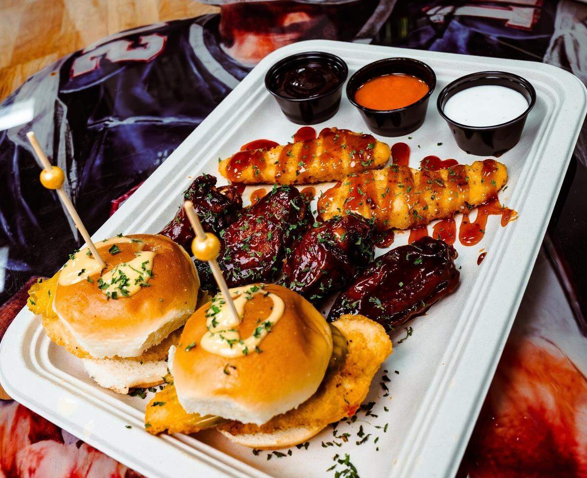 A white platter features a sampler of chicken sliders, glazed wings, and breaded tenders drizzled with sauce. Three small cups of dipping sauces—BBQ, buffalo, and ranch—sit at the top of the tray.