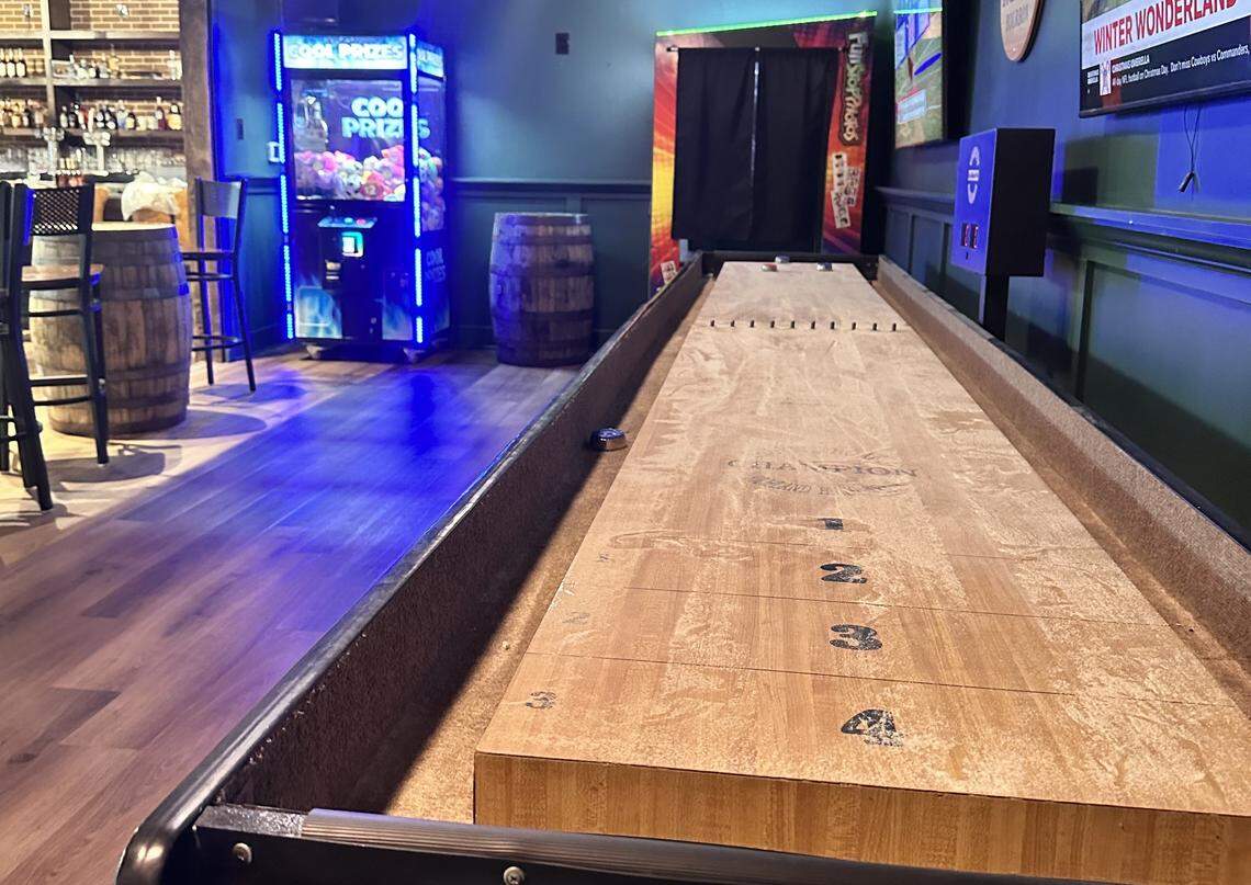 A close-up view of a long wooden shuffleboard table marked with scoring zones (1 through 4). In the background, a “Cool Prizes” claw machine glows with blue LED lights next to a wooden barrel and bar seating.
