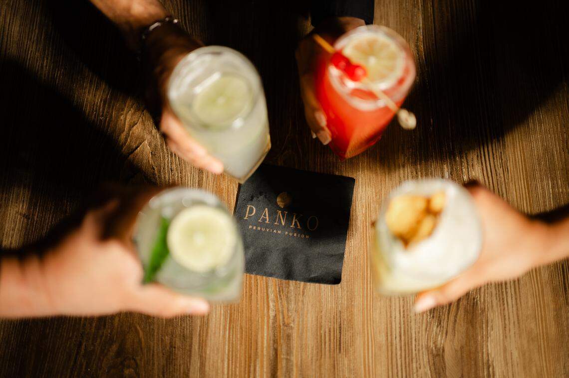Four hands holding assorted cold beverages are shown from an overhead perspective, creating a circular toast over a wooden table. The drinks, which include a red one garnished with a cherry and a slice of lime, frame a dark cocktail napkin featuring the PANKO Peruvian Fusion logo.