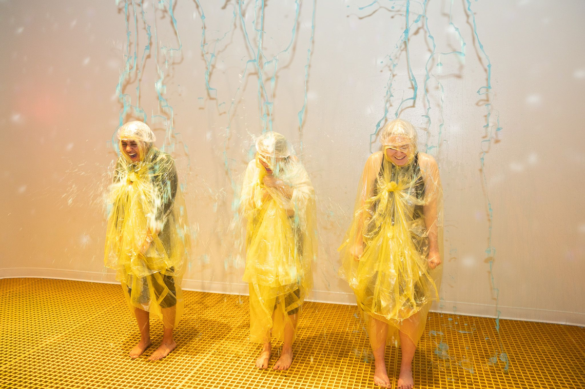 Three people wearing yellow ponchos and shower caps stand side-by-side as blue liquid streams down from above like a colorful indoor rain. They are laughing and bracing themselves while standing on a yellow grid floor against a plain white wall.