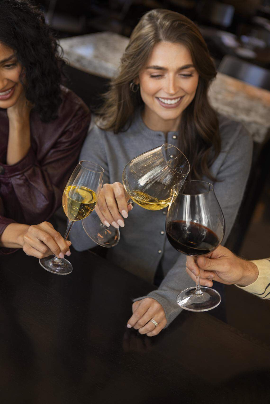 Three people are gathered around a dark wooden table, smiling as they clink wine glasses in a celebratory toast. The group is enjoying a variety of beverages, including a glass of red wine, white wine, and a flute of sparkling wine.