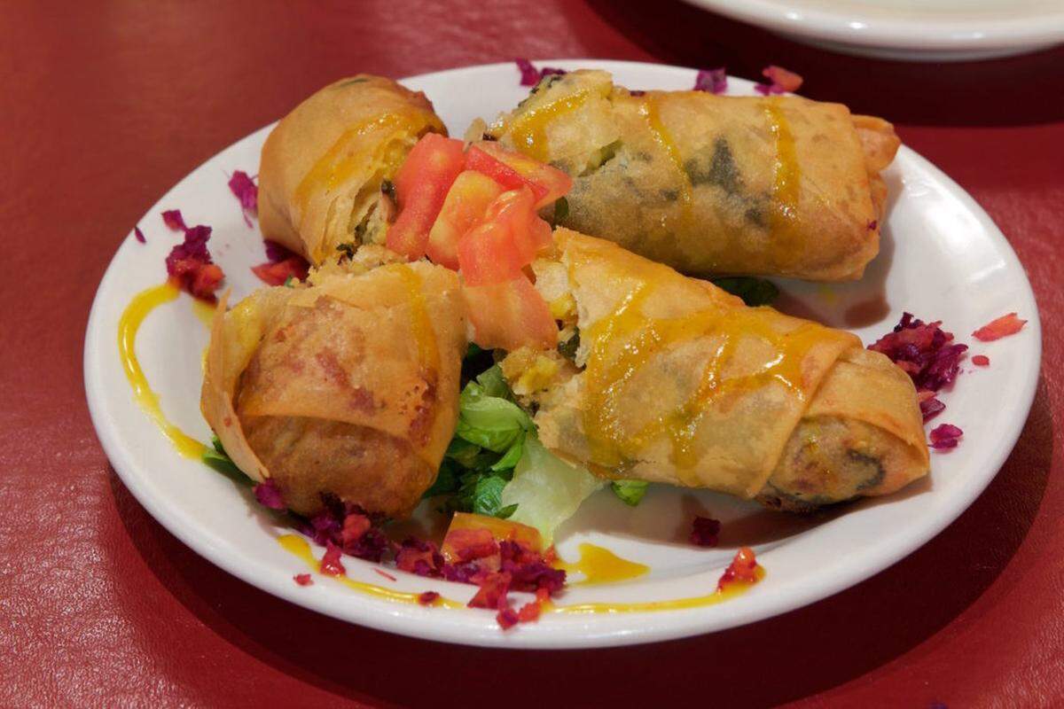 A close-up shot of a “Soul Roll” appetizer from Mert’s Heart and Soul, served on a white plate against a red background. Four crispy, golden-brown spring rolls are arranged on a bed of shredded lettuce. They are drizzled with a light yellow sauce and garnished with diced red tomatoes and scattered purple and red finely chopped ingredients, possibly red cabbage or beet. One roll in the foreground is slightly broken open, revealing its savory filling.