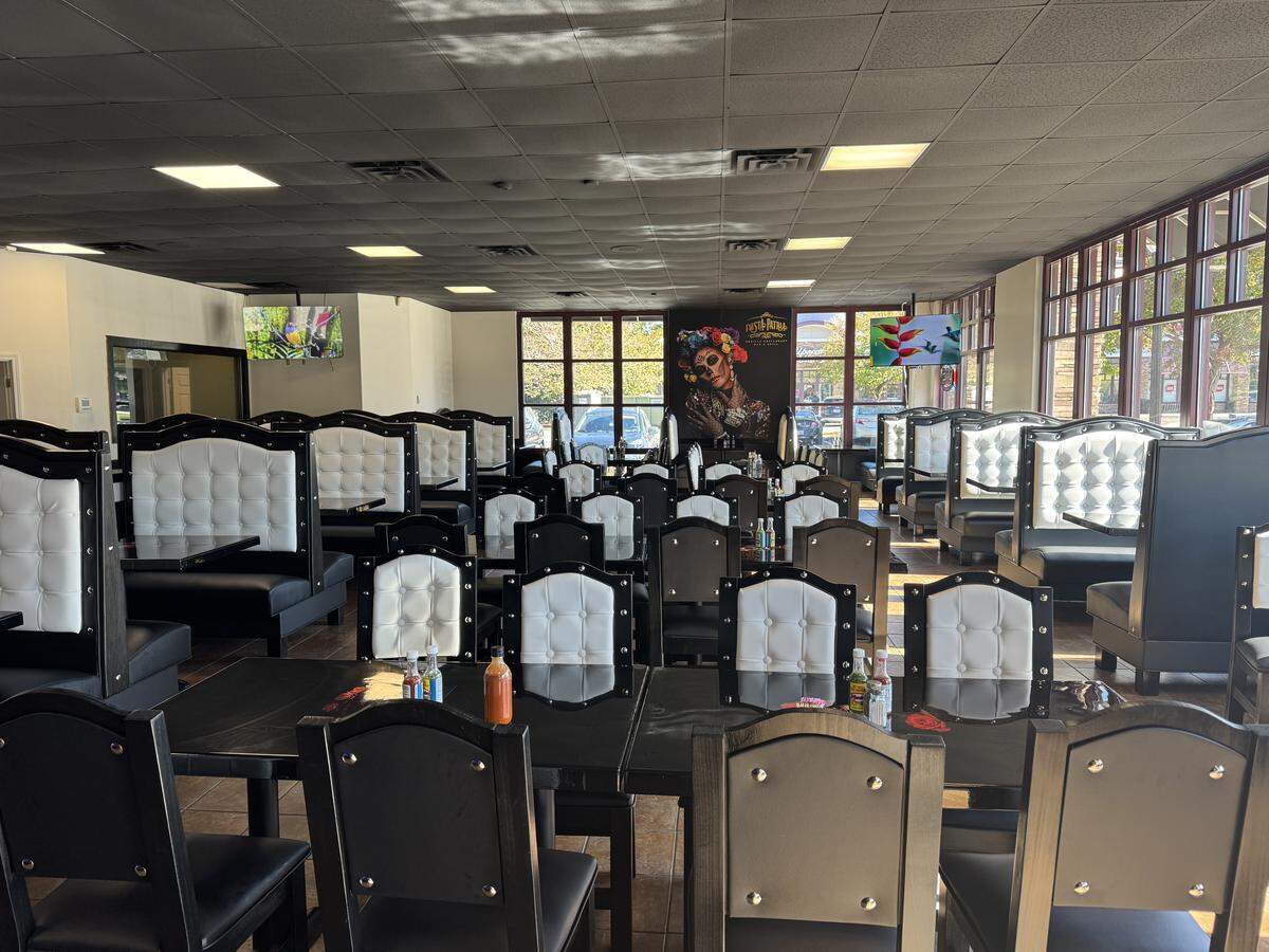 This is an interior shot of the Fiesta Patria Mexican restaurant dining room, showing rows of tables with matching black chairs and high-backed black booths with tufted white upholstery. Large windows line the back wall, and a colorful mural featuring a Día de Muertos theme is visible in the center.