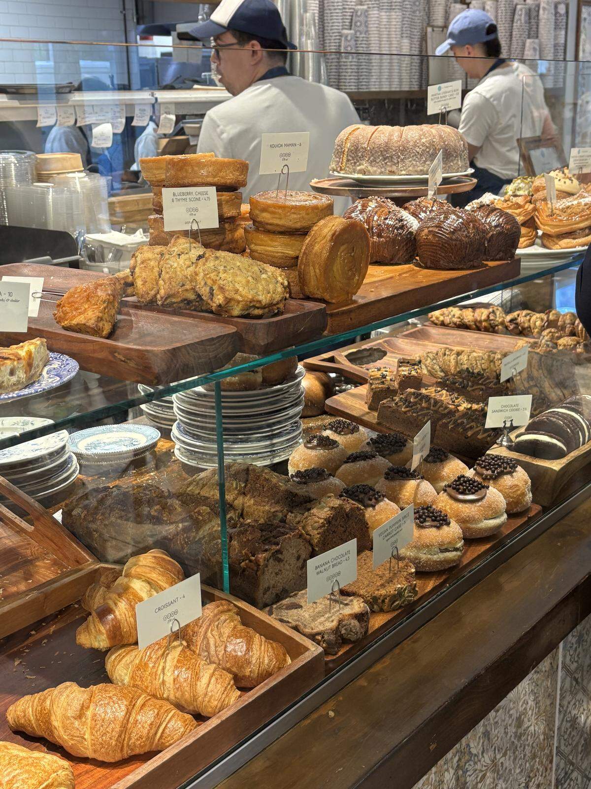 A glass display case is filled with an assortment of fresh pastries, including golden croissants, chocolate-topped donuts, and thick slices of banana bread. Behind the counter, bakery staff in white shirts and blue caps work in a bright, busy kitchen.