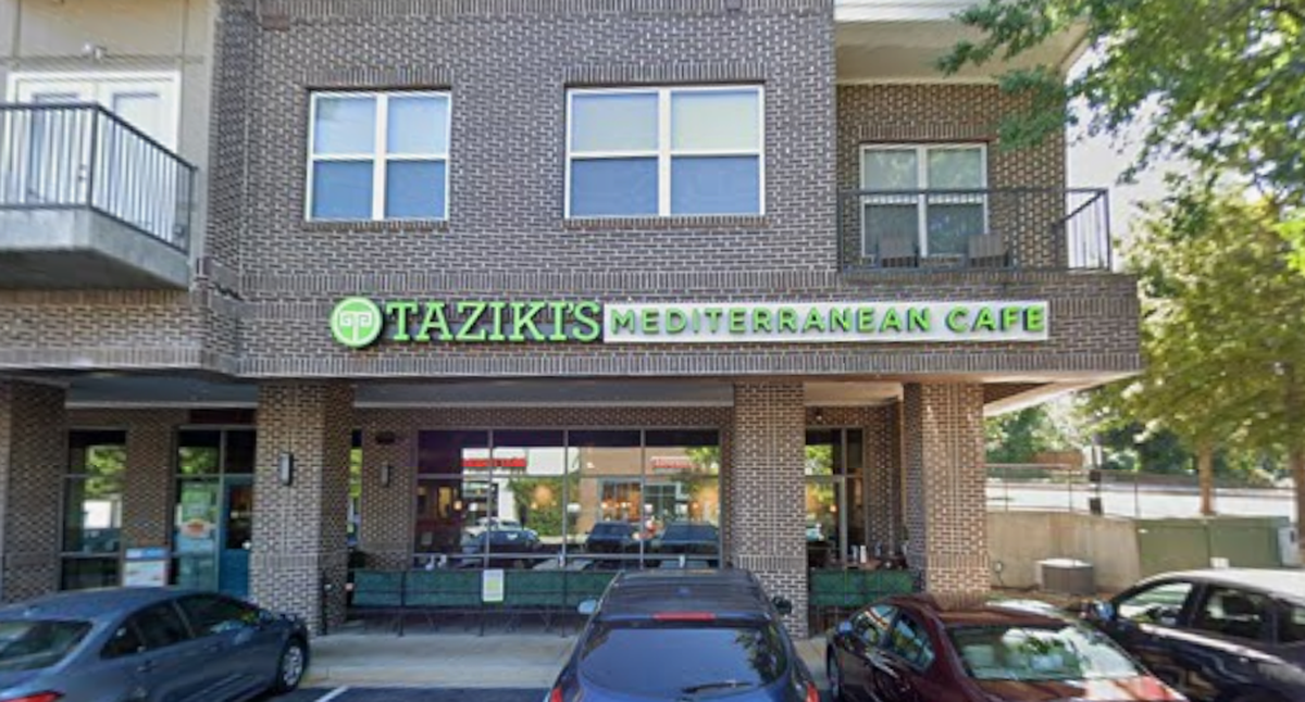A street-level photo shows the exterior of a Mediterranean restaurant named TAZIKI’S MEDITERRANEAN CAFE, which occupies the ground floor of a modern, multi-story brick building. Several cars are parked in the small parking lot directly in front of the storefront.