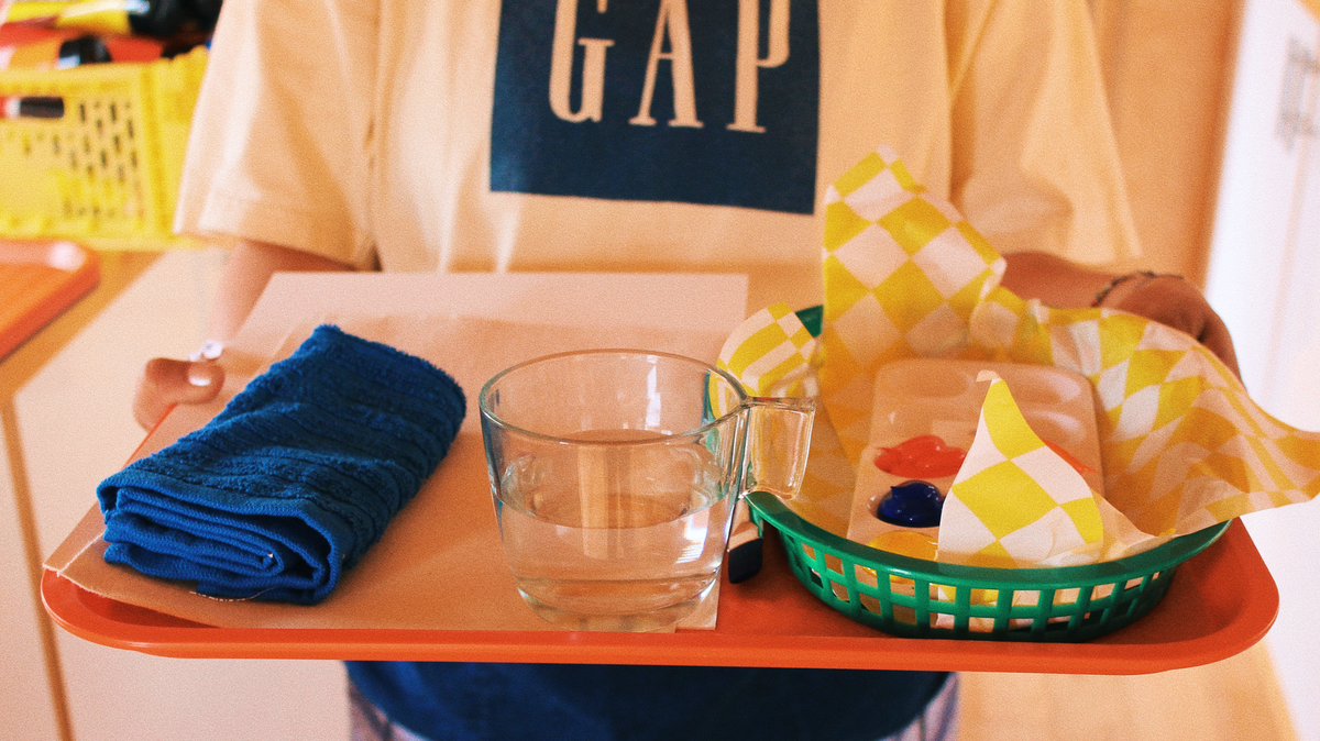A person wearing a Gap T-shirt holds an orange tray. On the tray are a small blue towel, a clear cup of water, and a green basket containing small paint tubes and a checkered cloth.