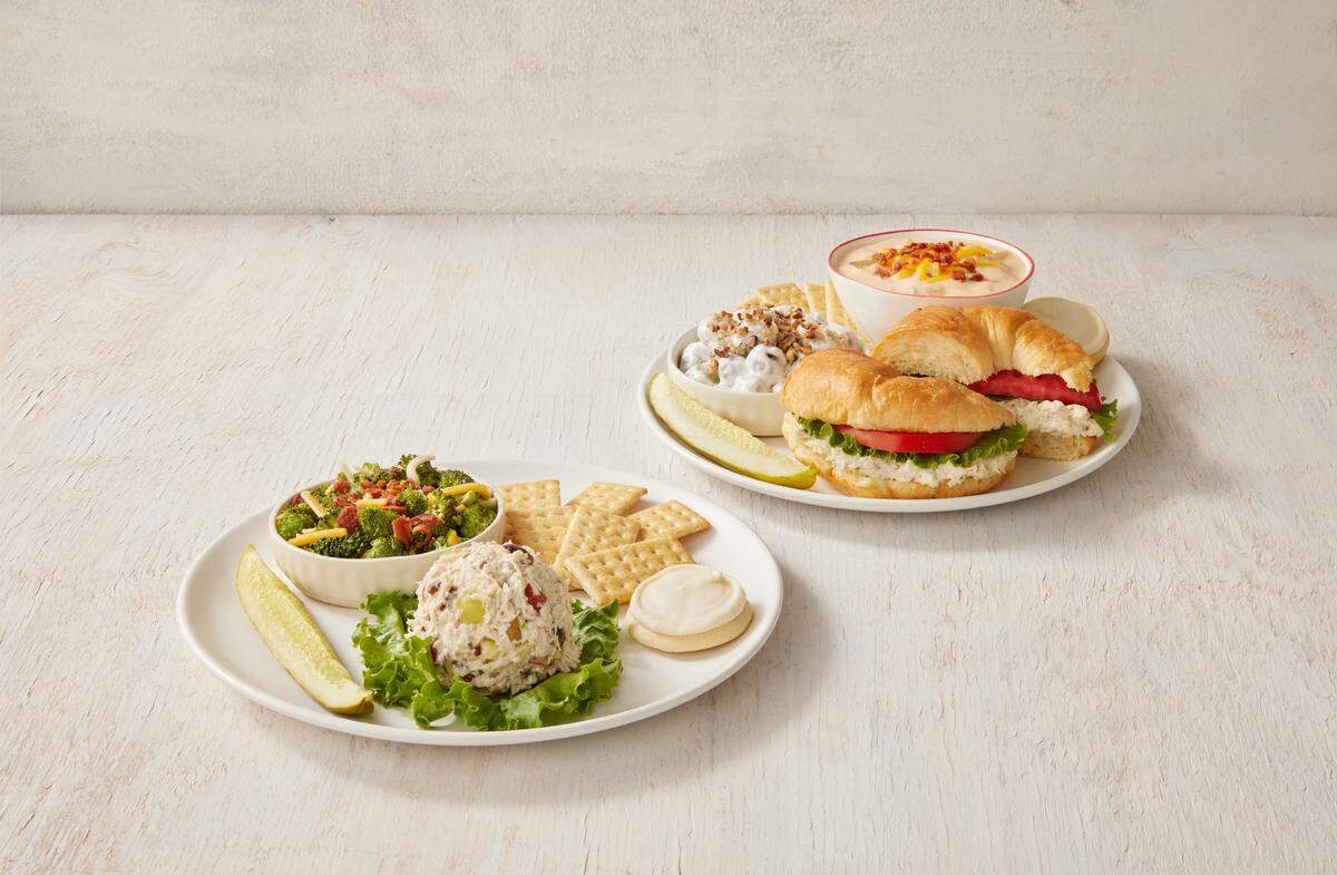 Two white plates are set against a light background, showcasing different meal combinations like a chicken salad scoop with broccoli salad and a croissant sandwich with potato soup. Both plates are garnished with a signature pickle spear and a frosted sugar cookie.