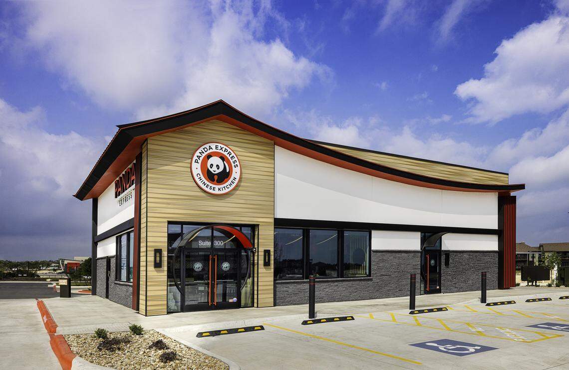 The exterior of a modern Panda Express restaurant features a distinctive sweeping roofline and a mix of light wood and stone textures. The building sits under a bright blue sky with its iconic panda logo prominently displayed above the glass entrance.