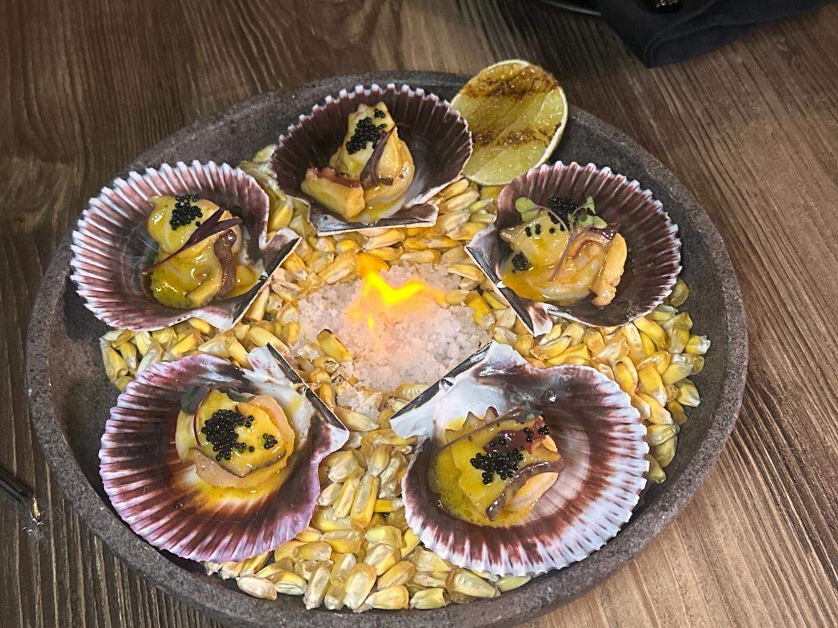 A dramatic presentation of scallops served in their shells arranged around a central mound of salt that is flaming. The seafood dish is set atop a bed of yellow corn kernels, garnished with black caviar, and includes a wedge of charred lime.