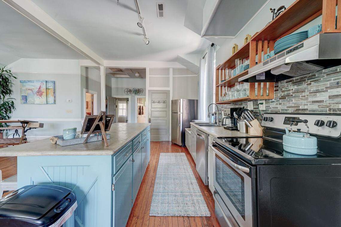 A bright kitchen showcases a large teal island with a white countertop and industrial metal stools. Behind it, a modern black stove sits against a multi-toned grey tile backsplash, complemented by open wooden shelving filled with blue and clear glassware. A long, light blue runner rug stretches across the hardwood floor toward a stainless steel refrigerator.