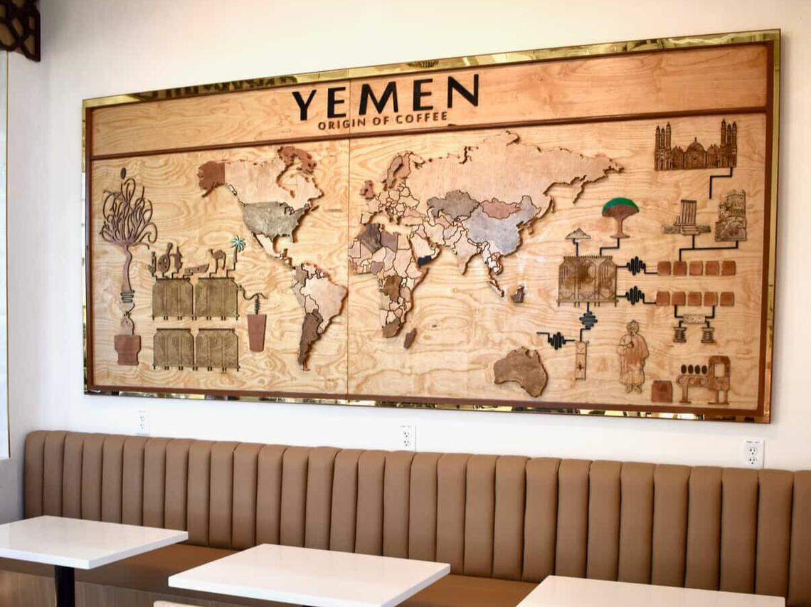 A large, horizontal wooden mural depicting a world map is mounted above a long, tan upholstered bench in a coffee shop. The map emphasizes “YEMEN: ORIGIN OF COFFEE” and features detailed wood carvings illustrating the coffee supply chain and significant landmarks.