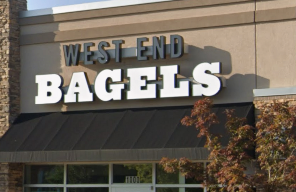 A storefront features a large sign reading “WEST END BAGELS” in bold block letters above a dark fabric awning. The words “WEST END” are written in smaller black text stacked directly above the larger, white word “BAGELS.” The exterior building blends tan stucco with stacked stone accents, and the unit number “1000” is visible on the glass window below.